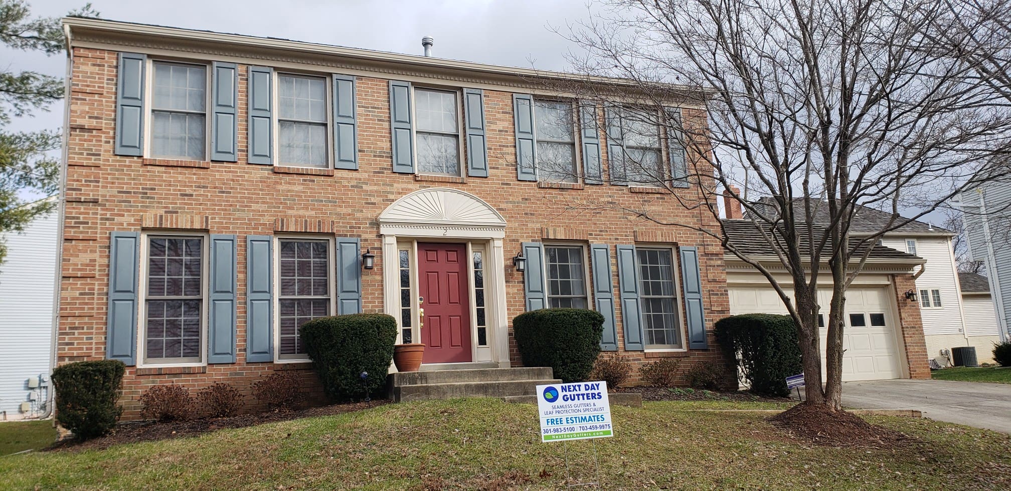 Brick colonial with blue shutters, a red front door, white gutters and a Next Day Gutters yard sign