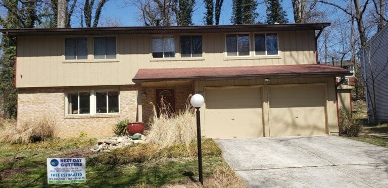 Two-story house with tan siding over a brick lower level and a Next Day Gutters yard sign