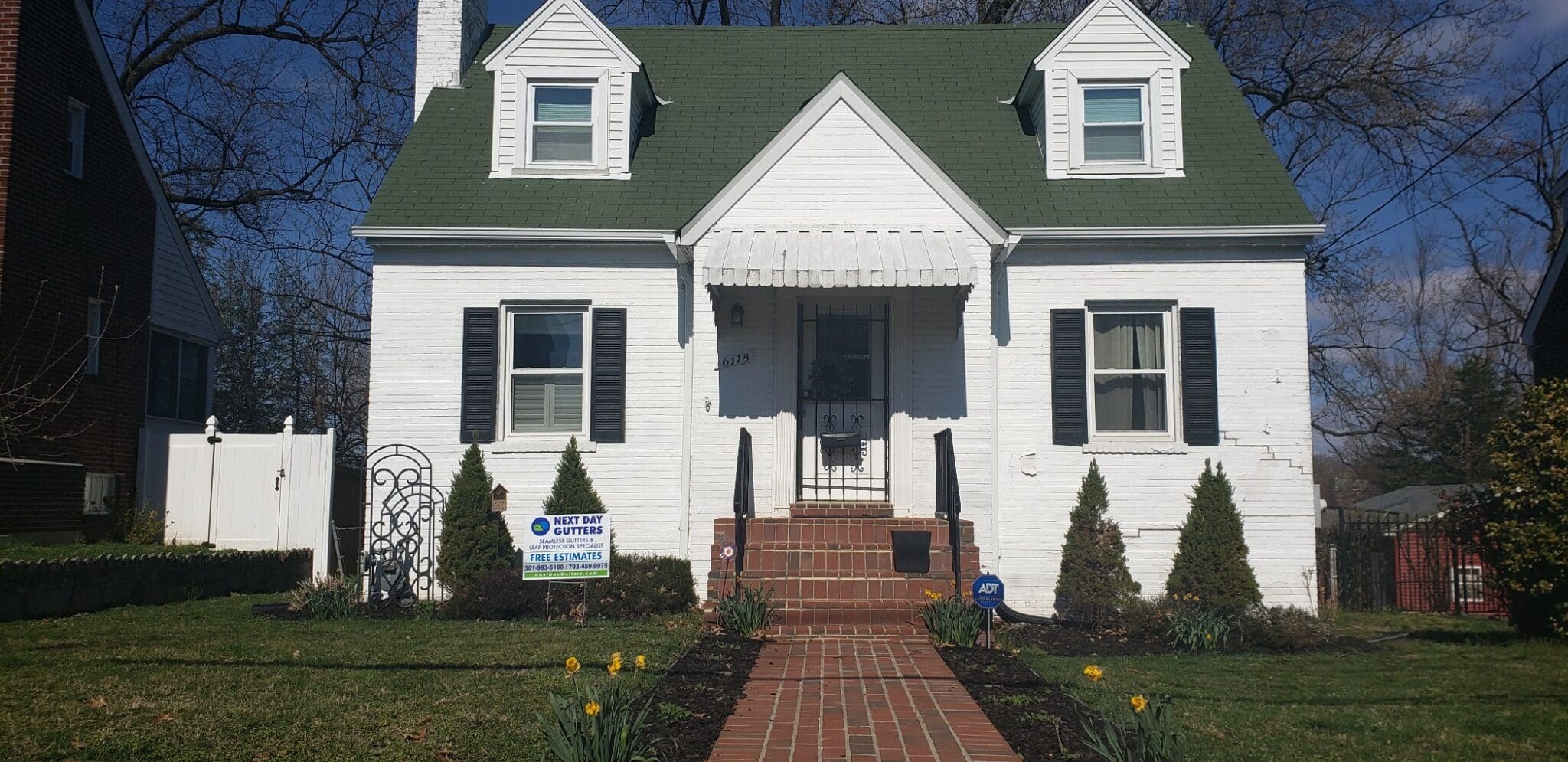 White painted-brick Cape Cod with black shutters, a green roof and a Next Day Gutters yard sign