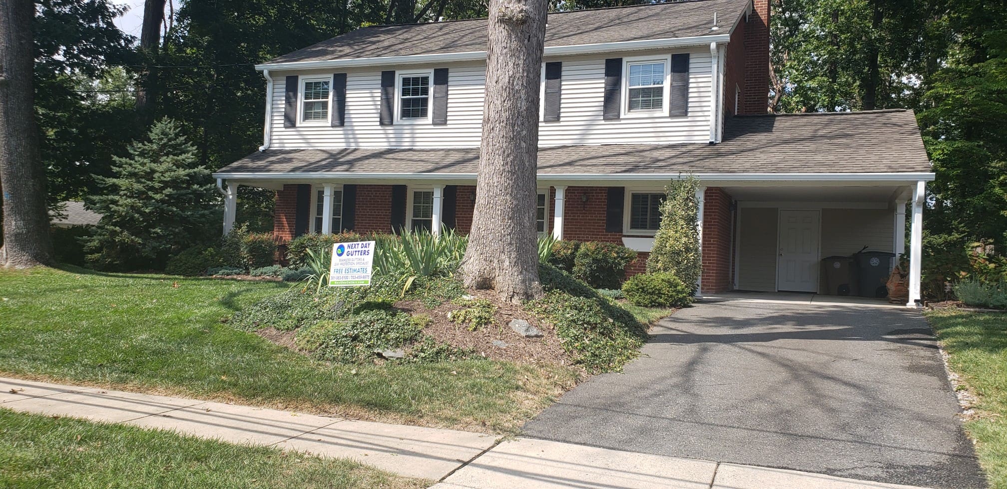 White two-story colonial with a brick base, black shutters, a carport and a large tree in the front yard