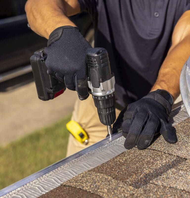 Installer in work gloves screwing a micro-mesh gutter guard panel onto a gutter with a cordless drill
