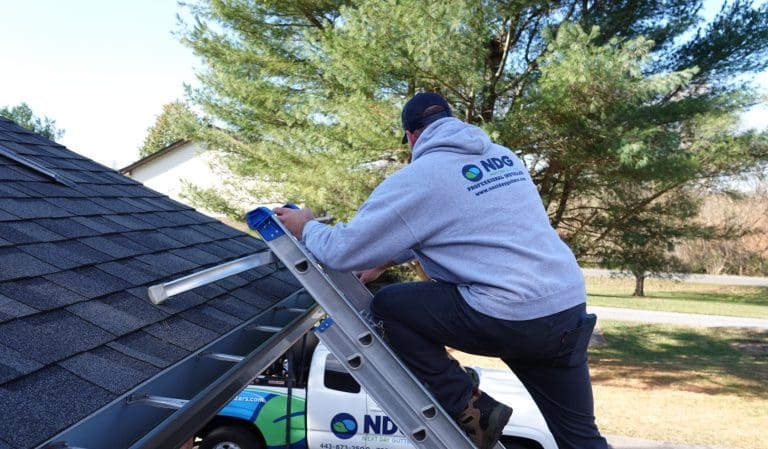Next Day Gutters installer on a ladder at the gutter line, with a company truck in the driveway