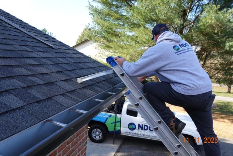 Next Day Gutters installer in a company hoodie climbing a ladder to a shingle roof, with the company truck in the driveway