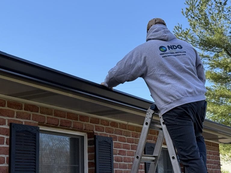 Next Day Gutters installer on a ladder working at the roof edge of a brick house
