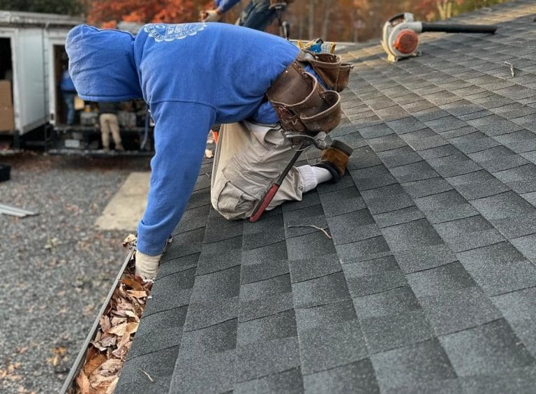Installer kneeling on a shingle roof fitting a gutter guard into the gutter